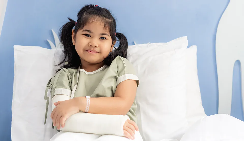 A child in a hospital bed wearing a cast on their right arm, looking at the camera.