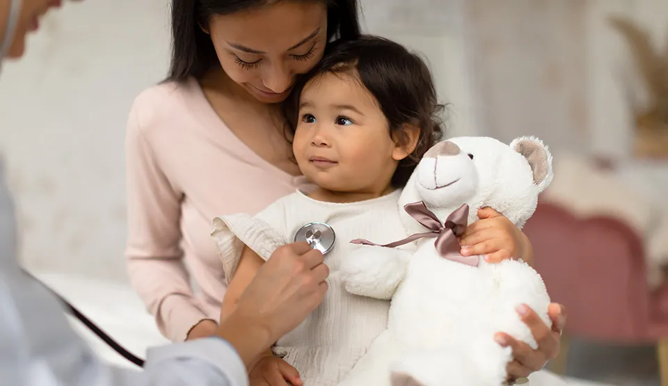 A doctor using a stethoscope on a baby holding a teddy bear.
