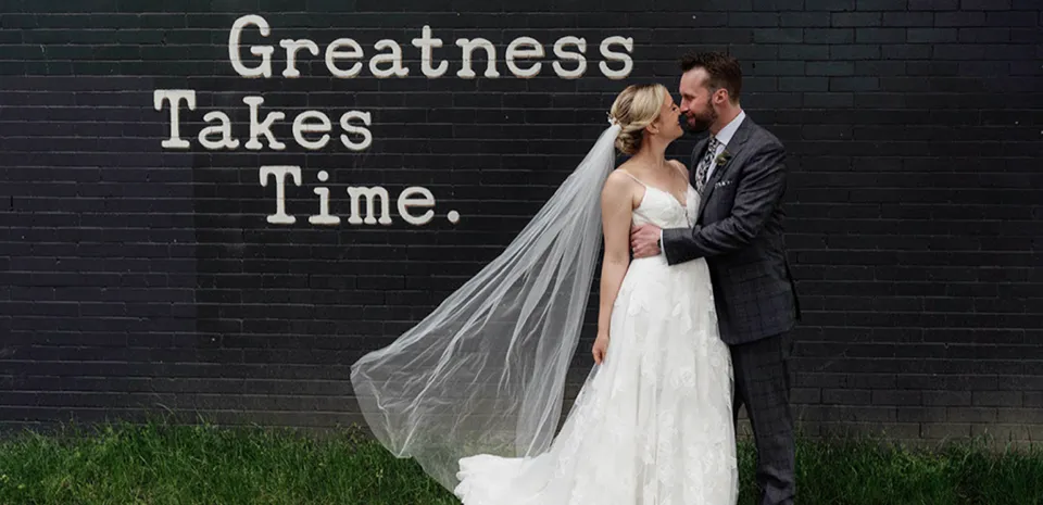 Husband and wife pose in an embrace for an outdoor wedding photo in front of a black, brick wall with Greatness Takes Time painted on it.