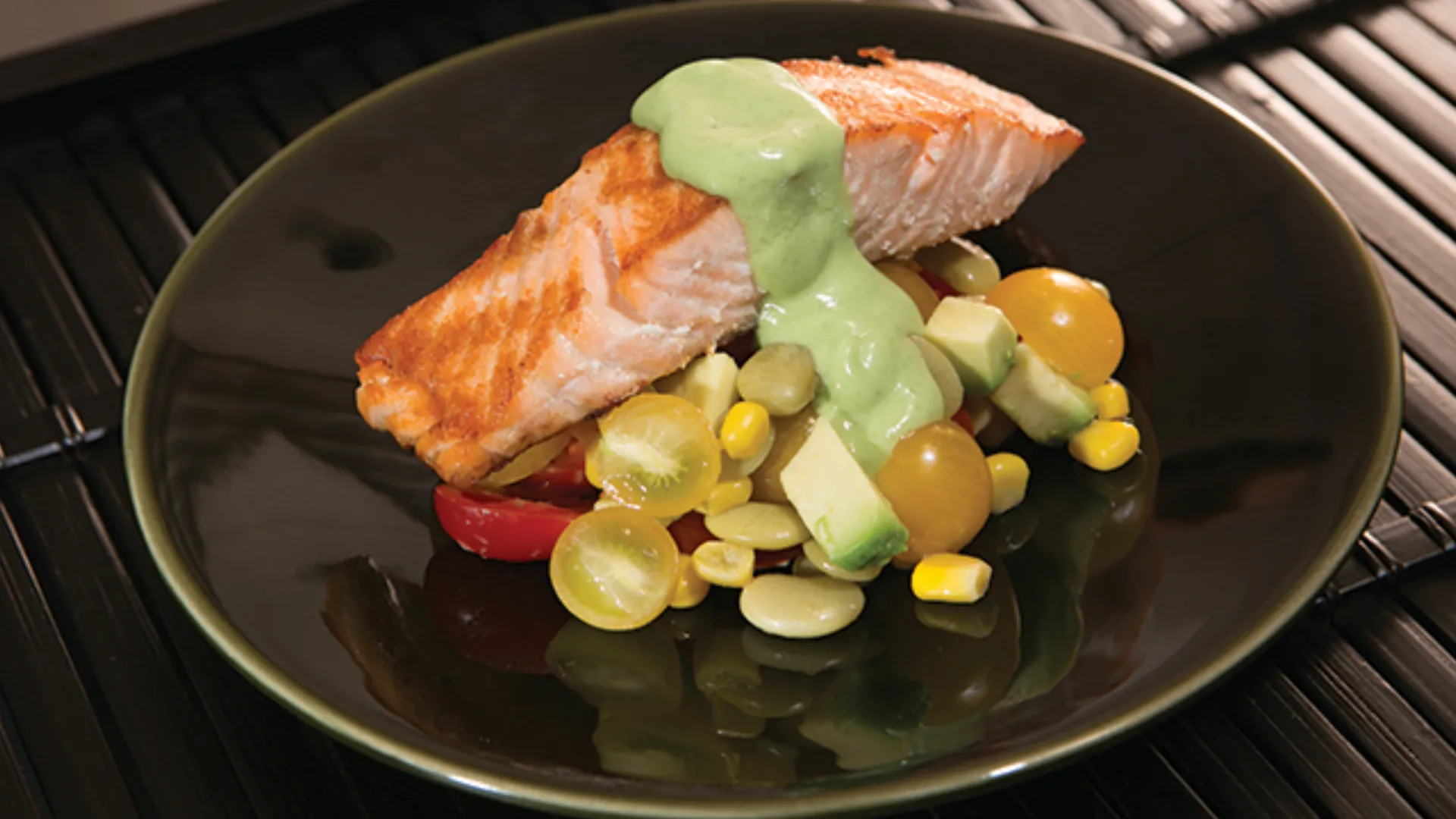 A piece of salmon with a green avocado sauce is next to a cherry tomato and corn salad, all on a black plate.