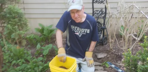 Peter Moniz kneels in a garden while using a yellow bucket.