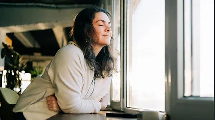 A woman sits at a window, eyes closed, taking a moment of mindfulness.