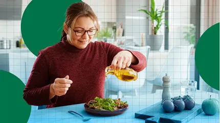 A woman pours oil over a salad while sitting at the kitchen table.