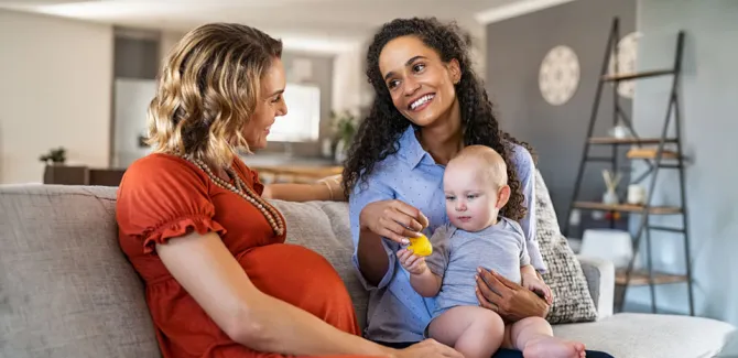 two women sitting on a couch looking at each other with a baby