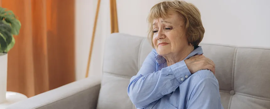 Woman sitting on a couch holding left shoulder wondering if her pain could mean a heart attack