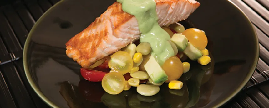 A piece of salmon with a green avocado sauce is next to a cherry tomato and corn salad, all on a black plate.